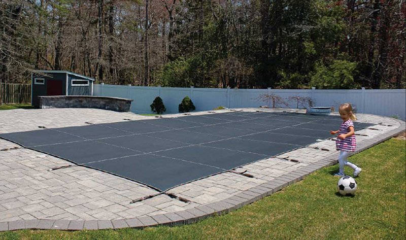 Gray mesh safety cover on a swimming pool with a child playing soccer next to it